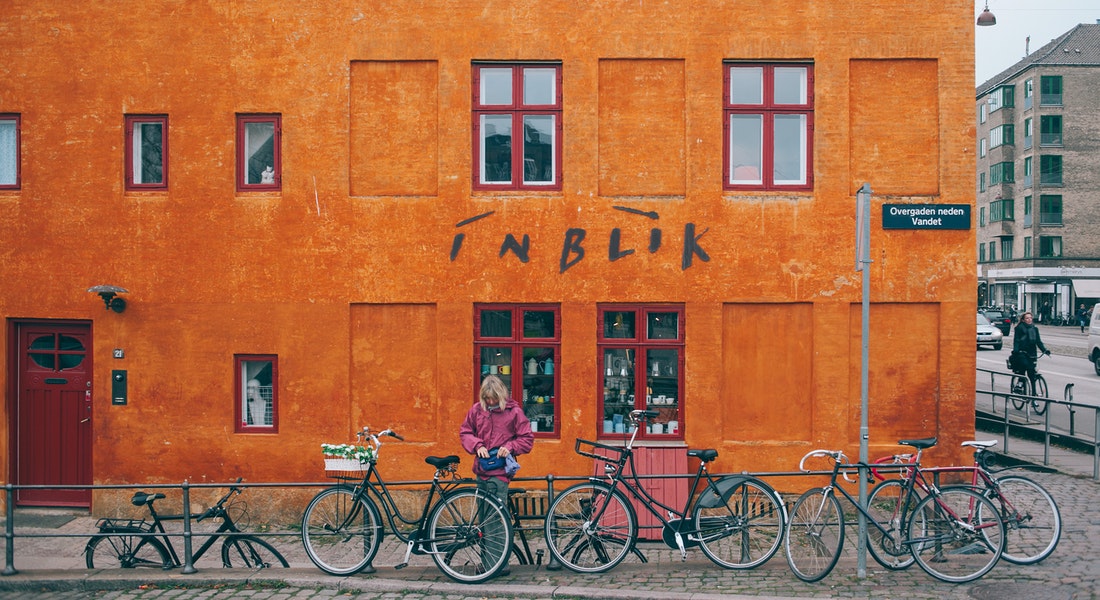 Female student standing with her bike in front of orange wall on building in Copenhagen showing how it is to be living in Copenhagen